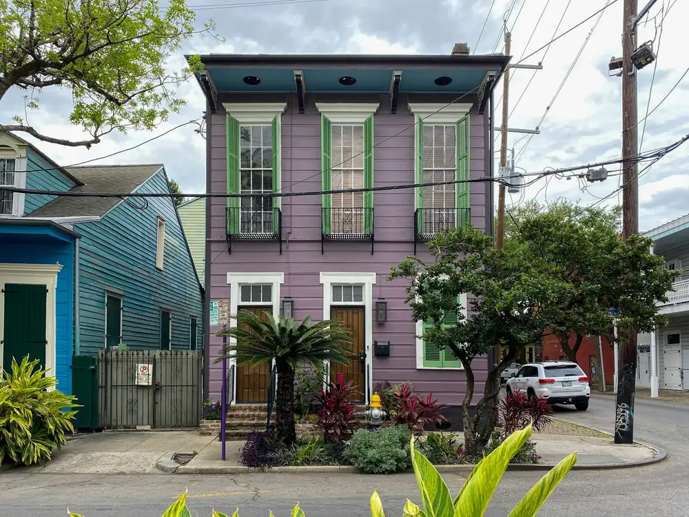 A purple two-story house with two front doors, green shutters and small iron balconies on the upper floor, on a street corner.