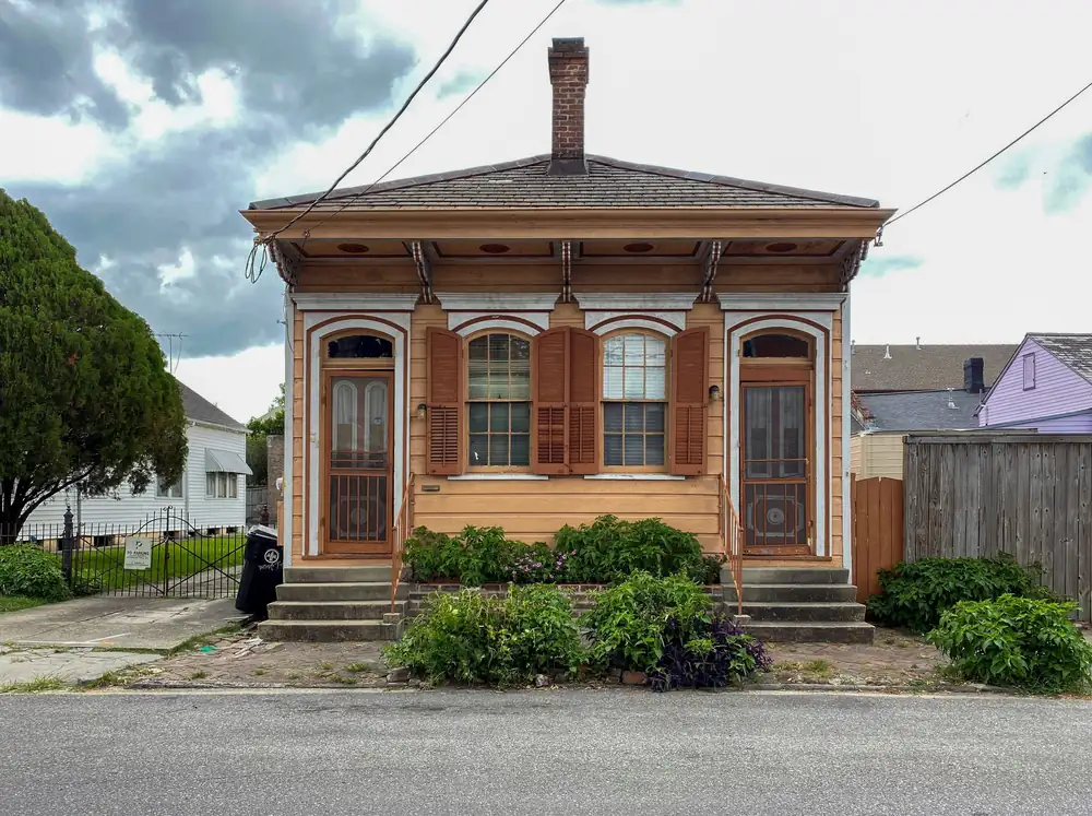 A tan one-story house with a hipped roof, a brick chimney and two front doors under a bracketed overhang, on a low raised base with a few steps at each door.