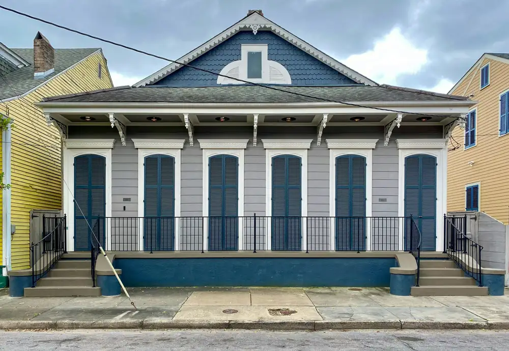 A gray double shotgun house with blue shutters and two front doors, a raised porch with an iron railing on a blue painted masonry base, and steps at each end.
