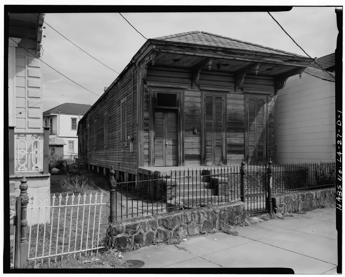 A historic black-and-white photograph of a long, narrow, weathered wooden house raised on piers behind an iron fence.