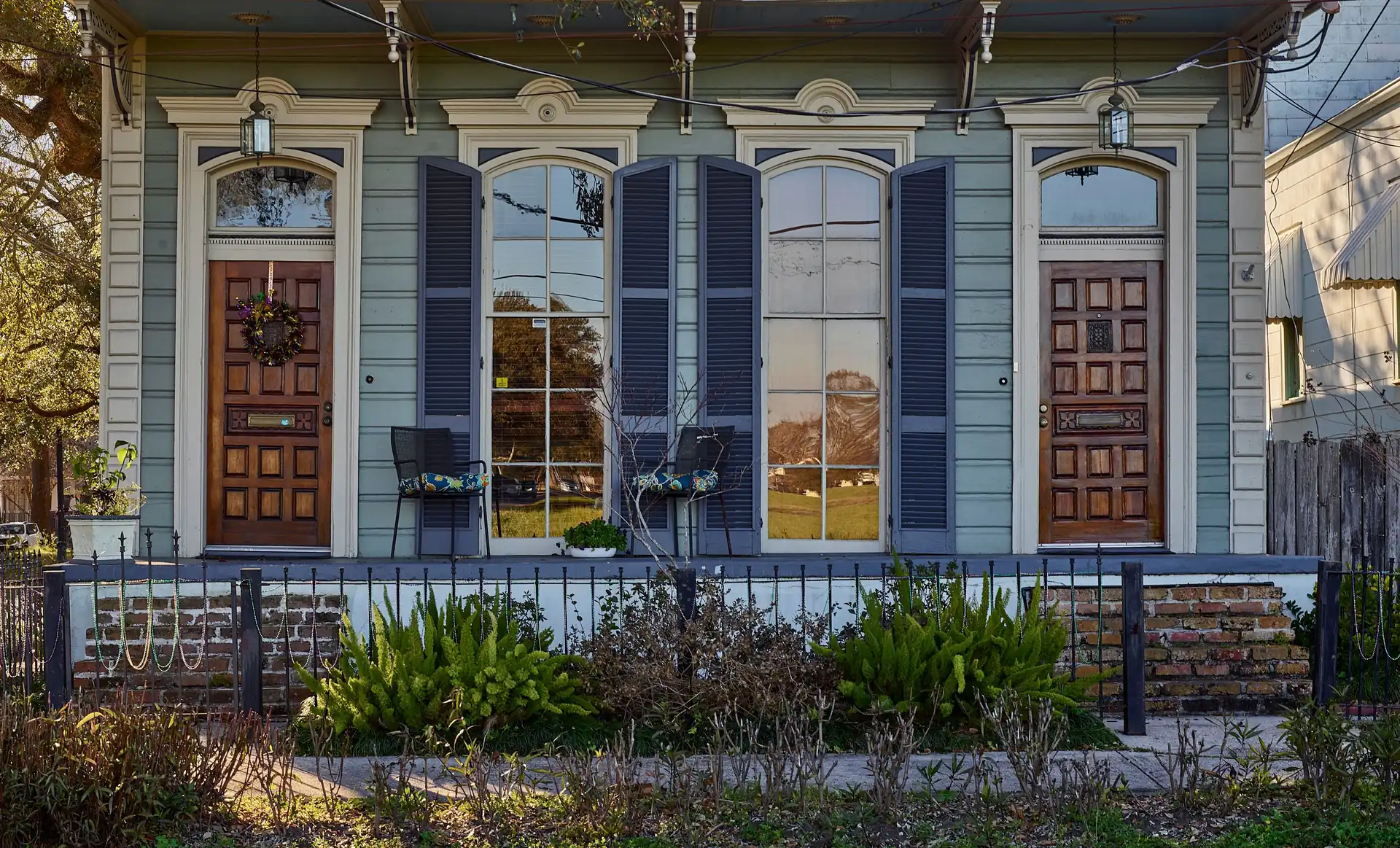 A raised New Orleans two-unit cottage with blue-gray shutters and two wooden front doors, on a brick base behind a low iron fence, in late-afternoon light.