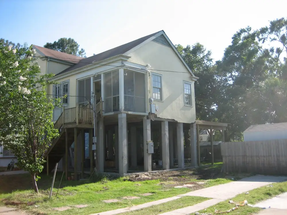A pale-yellow two-story house lifted high on tall concrete piers, with open space underneath and a timber stair on one side.