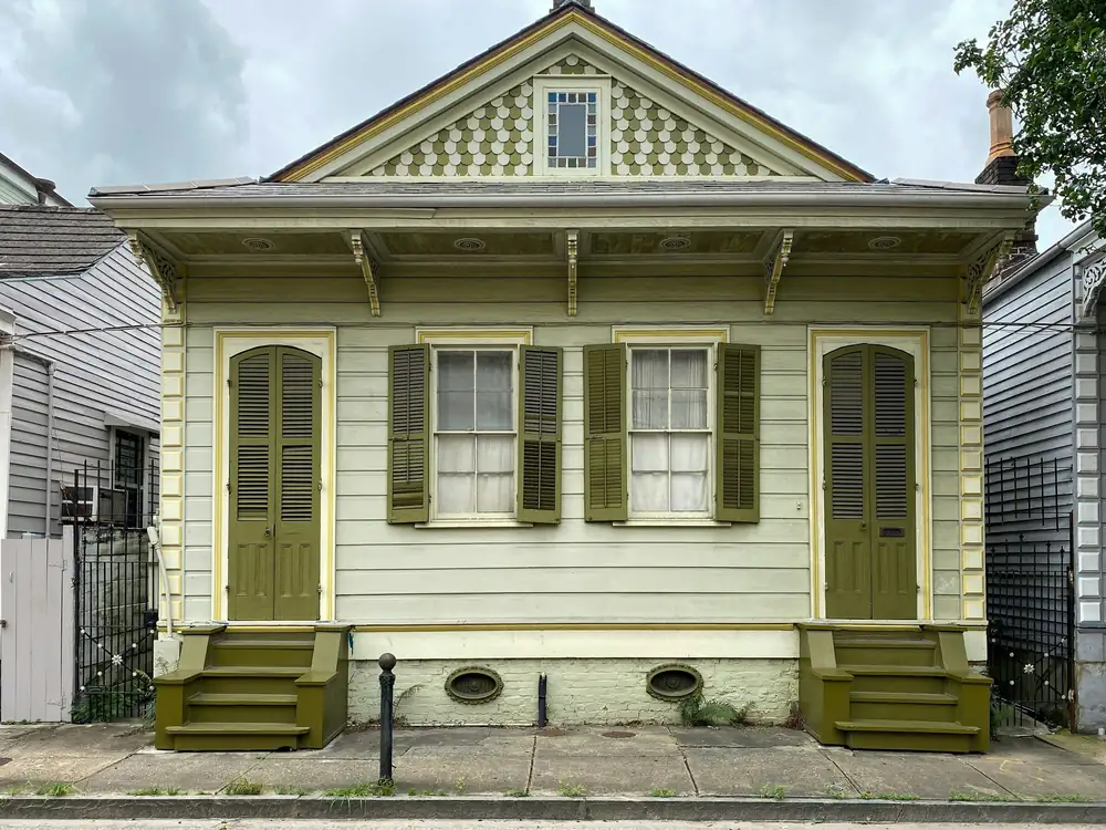 A pale-yellow wooden house with olive shutters and two front doors, on a raised stucco base with oval foundation vents.