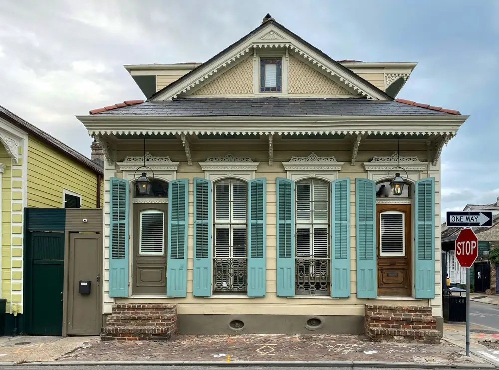 A cream house with teal shutters, ornate bracket trim and a lantern at each door, on a low raised base with two oval vents and brick steps at each end.